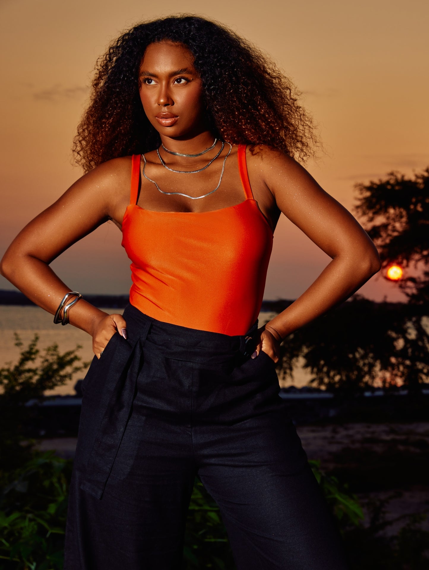 Modelo ao pôr-do-sol vestindo maiô elegante Laranja com Decote Reto e calça de linho preta.