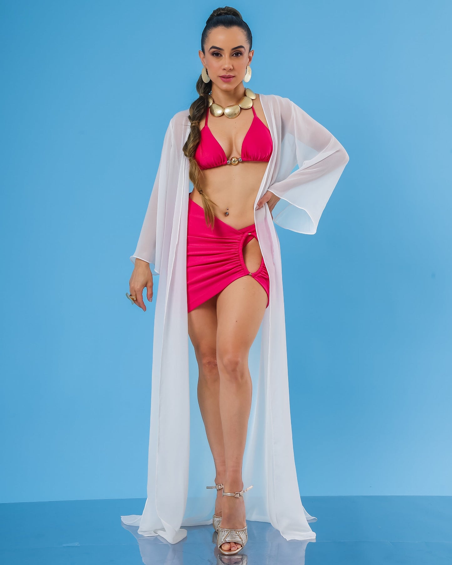 Modelo com look praia sofisticado e elegante composto por saida saia curta rosa pink, top biquini cortininha rosa e kimono branco transparente.