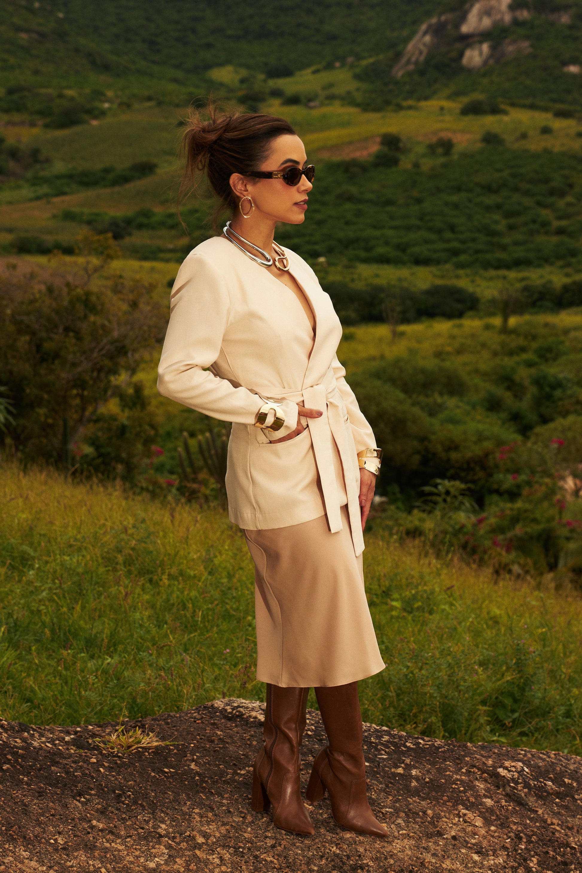 Mulher vestindo um blazer nude, saia midi e botas marrons, posicionada em uma colina, cercada por uma paisagem verde.