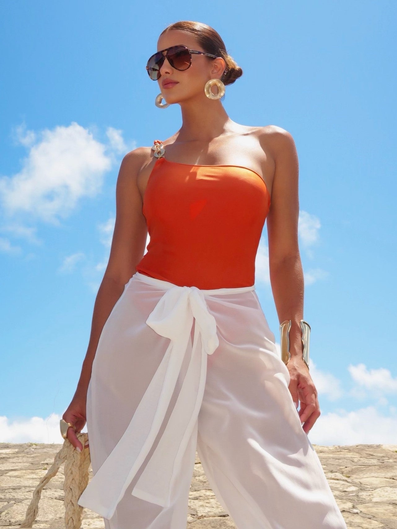 Modelo com look praia sofisticado, vestindo Maiô laranja elegante de um ombro só e saída de praia calça branca transparente.