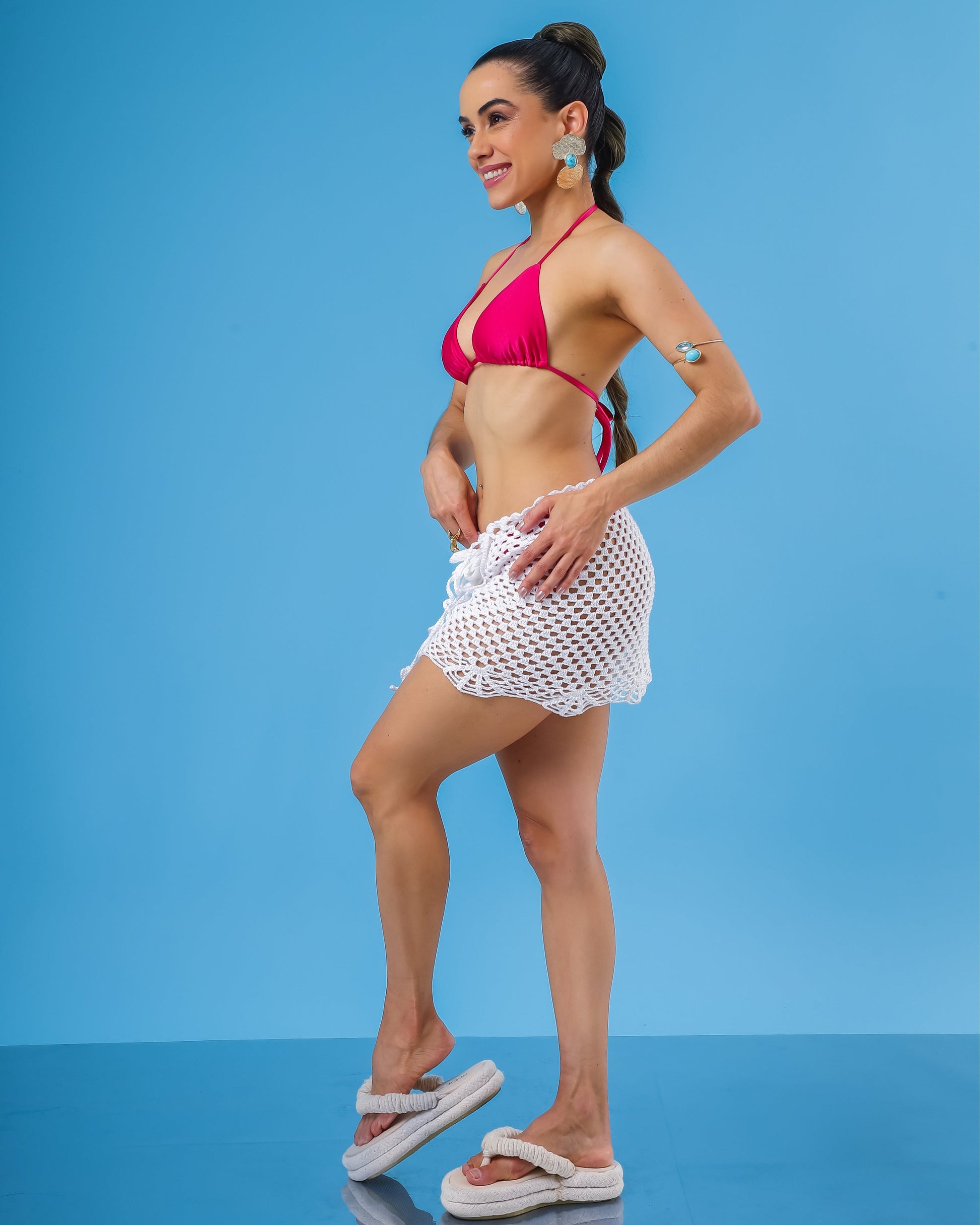 Modelo de lado vestindo saida de praia curta de croche branca, biquini cortininha rosa pink e sandália flat branca.